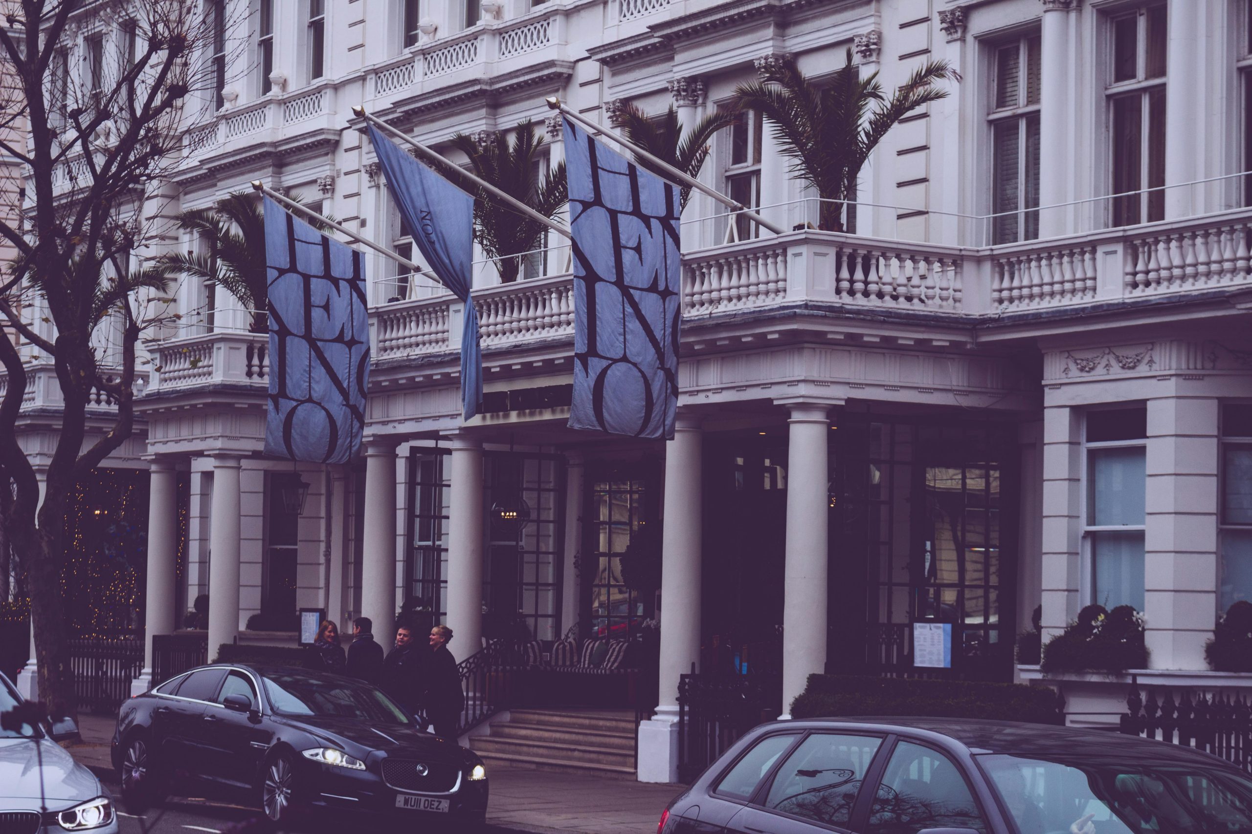 Street view of a hotel with flags, parked cars, and people outside, in a bustling urban environment.